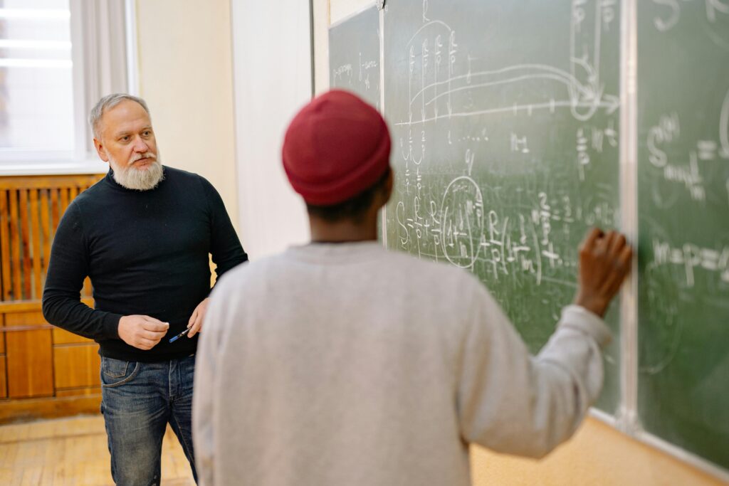 Educator assisting student during a math lesson at a blackboard in a classroom setting.